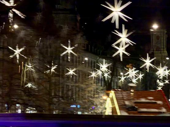 Eine nächtliche Szene mit dekorativen sternförmigen Lichtern, die über einer Straße hängen, beleuchteten Gebäuden und einer teilweise sichtbaren festlichen Struktur, wahrscheinlich einem Marktstand. Das Bild hat einen verschwommenen Effekt, was auf Bewegung hindeutet.