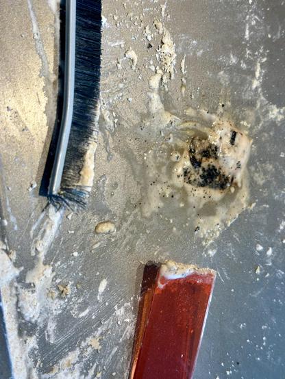 Close vote of stainless steel baking sheet with  thin hard black brush and hard red nylon small spatula. Baking soda residue all over pan. The black burned blob (BBB) is center left. It is sticking up 1/8” and it’s the size of a small pancake. It is moist from spraying hydrogen peroxide on it for a paste. Some of the blob is turning brown.