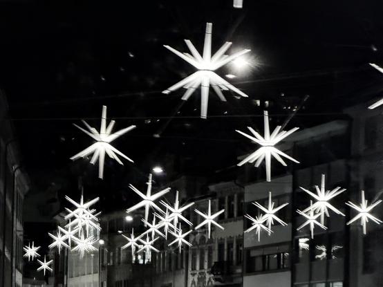 Eine Nachtszene mit hellen, sternförmigen Dekorationen, die über einer Straße hängen. Der Hintergrund zeigt Gebäude, die von Straßenlaternen und Reflexionen beleuchtet werden.