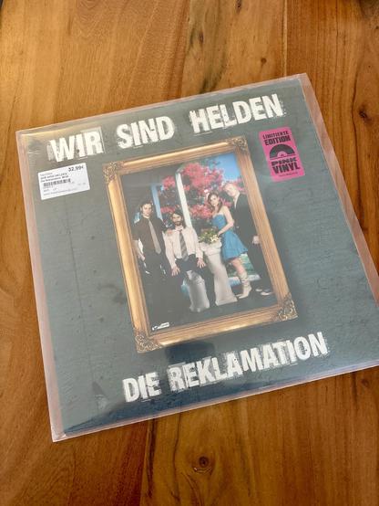 Ein Vinyl-Cover mit dem Titel "Die Reklamation" von Wir Sind Helden. Es zeigt eine Gruppe von vier Personen, die in einem gerahmten Porträt vor einem Hintergrund von Blumen posieren. Das Cover wird auf einer Holzoberfläche präsentiert, mit einem Schild, das darauf hinweist, dass es eine limitierte pinke Vinyl ist.