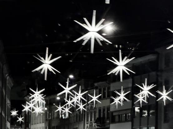 Eine Nachtszene mit hellen, sternförmigen Dekorationen, die über einer Straße hängen. Der Hintergrund zeigt Gebäude, die von Straßenlaternen und Reflexionen beleuchtet werden.
