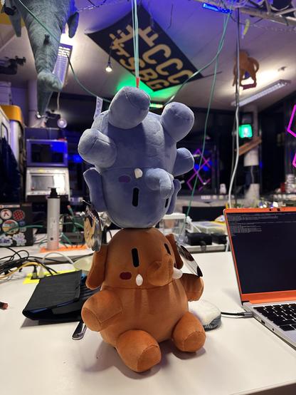 A blue and a brown plushtodon plushies are stacked on a white table. The background features various items and colorful lighting, a blåhaj and a cccsbg flag.
