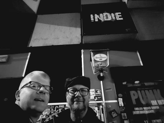 Zwei Männer stehen vor einer Kneipe, über deren Tür ein Schild mit der Aufschrift "Indie" hängt.
