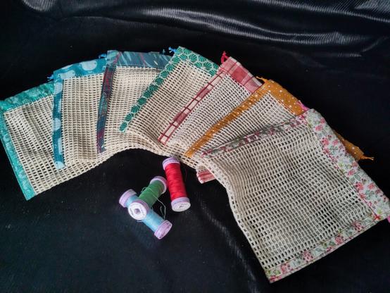 7 sacs a savon en éventail sur un fond noir. ce soit des petit sacs carré avec un fillet au centre et des bouts de tissus colorés sur les bords. il y a quelques bobines de fil en dessous