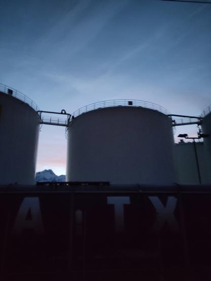 Foto. Industriegebiet mit zylinderförmigen Bauwerken, davor die Lettern (G)ATX, im Hintergrund ein Berg.