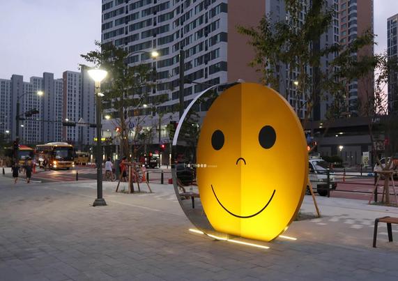 A large illuminated smiley face sculpture stands on a busy street, surrounded by buildings and pedestrians during twilight.