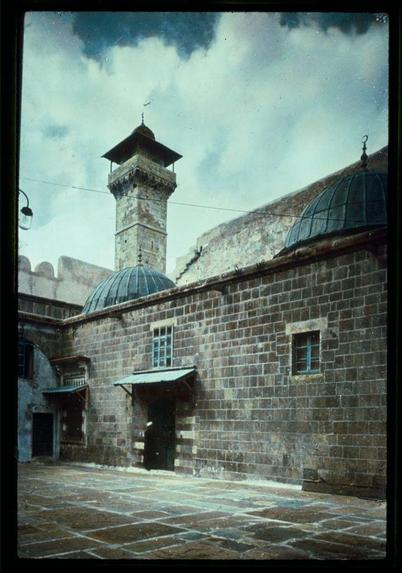 The image depicts an architectural structure with a blend of historical and religious significance. In the foreground, there is an open courtyard paved with stone slabs, showcasing signs of wear which suggests age or heavy use over time.

Two prominent domed structures rise from either end of what appears to be a large rectangular building constructed with dark brown brickwork. These domes have iron grills on their top and small windows at the base level, possibly serving as ventilation for storage below or simply decorative elements typical in Islamic architecture.

The central focus is an older stone minaret tower, featuring weathered stonework indicative of considerable age, likely centuries old. The design includes a pyramidal roof with a weathervane on top and smaller dome-like structures at the base, characteristic of many historic mosques or religious institutions found across the Middle East.

The background shows part of another stone structure with similar architectural style to that in the foreground, suggesting this could be within a walled complex like an old city's fortress. The sky is overcast with white clouds against a pale blue backdrop and there are modern street lamps visible on the left side, indicating contemporary maintenance or use of these historic sites.

The image appears aged as if taken several decades ago based on its graininess and color saturation; it carries historical weight, likely dati [...]