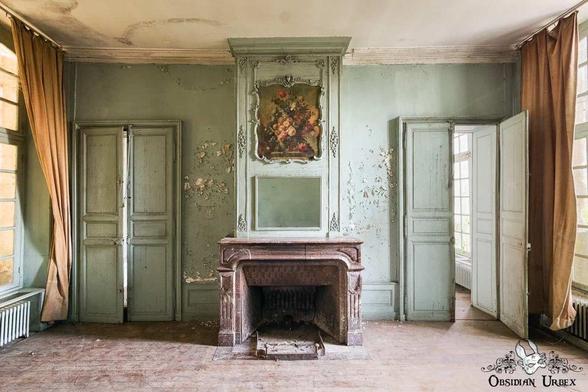 The room features a prominent marble fireplace and original, faded green paneling and doors.
The wallpaper is peeling, revealing floral patterns, and the floorboards are bare and worn.