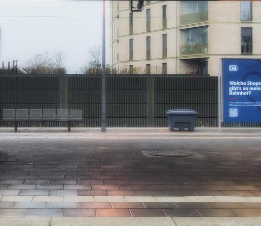 Szene auf einer leeren S-Bahn-Station. Alles ist rechteckig, auch das mehrstöckige Haus im Hintergrund. Der Himmel ist grau, der Bahnsteig nass. Das einzig farbige ist ein Plakat am rechten Rand, selbst das ist langweilig, weiße Schrift auf blauem Grund.
Scene at an empty suburban railway station. Everything is rectangular, including the multi-storey building in the background. The sky is grey, the platform wet. The only colour is a poster on the right-hand side, but even that is dull, with white lettering on a blue background.