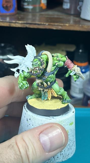 A green mean orc miniature on a sand base, blue tacked to a cork (between my fingers). He’s dressed in the bones of different dinosaurs, wearing a triceratops skull as a shoulderpad. There’s some body piercing (using giant teeth) and he’s really beefed up (and angry). The skin is painted in cold green tones (with warmer, more yellow tones over the shoulders). He’s also using a big avian skull as a punch dagger, tied to his left wrist. There’s a pink bit: a piece of flayed human skin (probably a face).