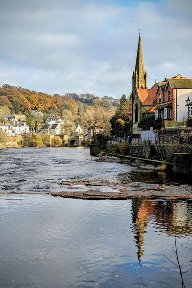 A photograph taken in late November of the River Dee flowing through the town of Llangollen, Wales. In the foreground, the river is low, exposing mossy rocks and autumn leaves. On the right bank stands the Llangollen Methodist Church with its tall, Gothic-style spire, nestled among historic town buildings. The opposite bank features white-washed houses and a stone bridge, backed by hills covered in deciduous trees displaying rich autumnal colours under a partly cloudy sky. The church and structures are clearly reflected in the calmer water closest to the viewer.