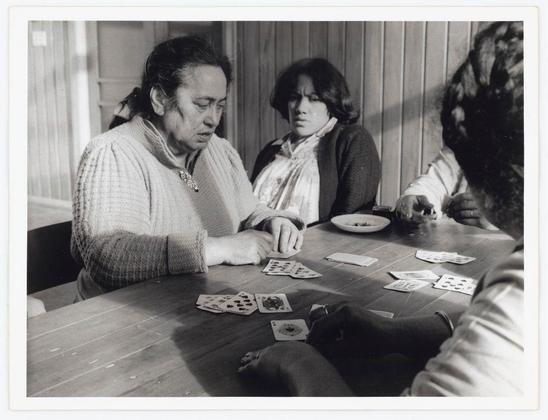 The black and white photograph captures a candid moment among individuals engaged in playing cards. Three people are seated at a table, with their attention focused on the game before them.

On the left side of the image is an older individual wearing a knitted sweater adorned with what appears to be a brooch or pendant around the neck area. This person's hair is dark and pulled back from the face, and they seem deeply engaged in examining cards spread out on the table.

In the center sits another participant, whose attire includes a cardigan over their clothing, paired with a scarf. Their expression suggests concentration as they look at their hand of cards.

On the right side, only partially visible is someone who appears to be reaching for or handling one of the playing cards scattered across the wooden surface. The individual's sleeve and part of what seems like dark hair are in view but not much else can be discerned due to the angle.

The table holds an assortment of card decks spread out with various faces up, indicating active gameplay. A bowl is also present on the tabletop near the center person, though its contents aren't clear from this perspective.

In summary, it's a snapshot that conveys a sense of social interaction and leisure through playing cards among what seems like friends or acquaintances in an indoor setting.