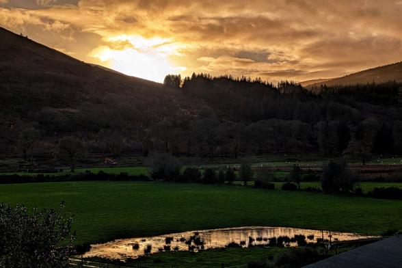 A dramatic landscape captured during golden hour, featuring a dark, silhouetted hill and thick tree line set against a sky bursting with bright orange and yellow light breaking through clouds. A lush green field stretches across the mid-ground, leading to a small, irregular body of water in the foreground that reflects the fiery colours of the sunset.