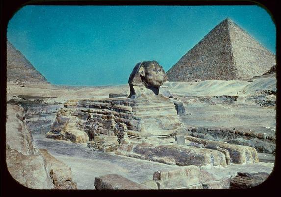 The image shows the iconic Great Sphinx of Giza, with its massive head and body composed of limestone. The face has a human-like expression and is highly detailed, suggesting it was carved from one large block. Behind the Sphinx are two towering pyramids known as the Pyramid of Khufu (also called the Great Pyramid) on the left side and possibly another pyramid to the right, which may be that of Khafre or Menkaure.

The ground appears sandy with patches of erosion and wear, indicative of an arid climate. The sky is a clear blue without any visible clouds. A slight vintage tint suggests this photo might have been taken during the mid-20th century.

This image captures one of Egypt's most famous landmarks and offers insight into ancient Egyptian architecture and sculpture techniques.