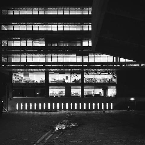 High contrast square monochrome photo of the University of Edinburgh main library building at night. 7 stories of almost regular lit windows, but a few rooms are not lit, and the top right is obscured by the bulk of the adjacent lecture theatre. In the centre foreground, a small puddle reflects some of the lights.