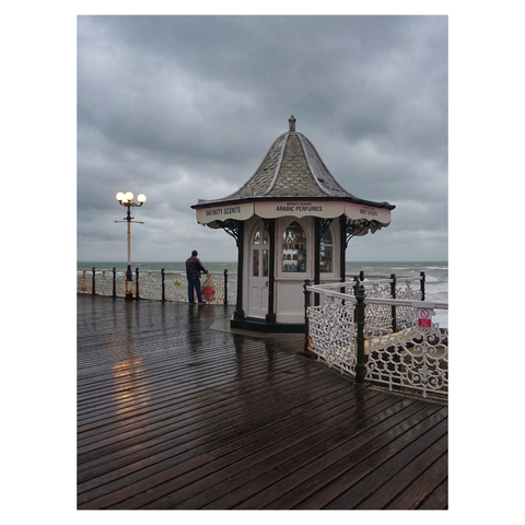 This is a portrait format colour image of a Victorian era hexagonal booth on a pier. The backdrop is stormy. An ornate lamppost is illuminated against the moody sky to the left and is reflected in the damp wooden slats of the pier. A man stands looking out to sea standing between the lamppost and the booth. Taken in Brighton with a Sony RX100.