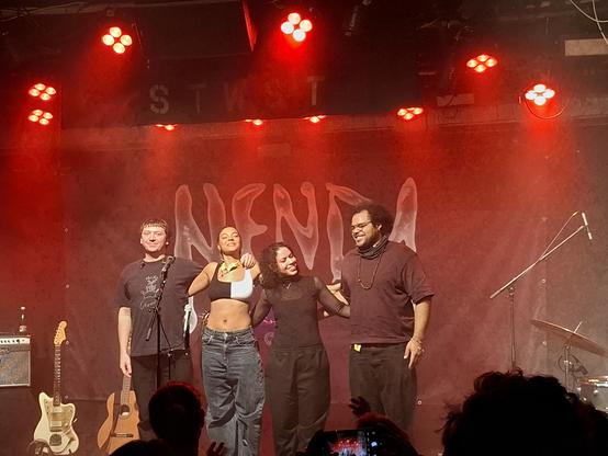 Abschlussfoto von Nenda mit Band die nebeneinder stehen. Rotes Licht kommt von oben. Im Hintergrund der Schriftzug "Nenda". Links sieht man zwei Gitarren stehen. Nenda trägt ein schwarz weißes bauchfreies Top. Die Band ist schwarz gekleidet