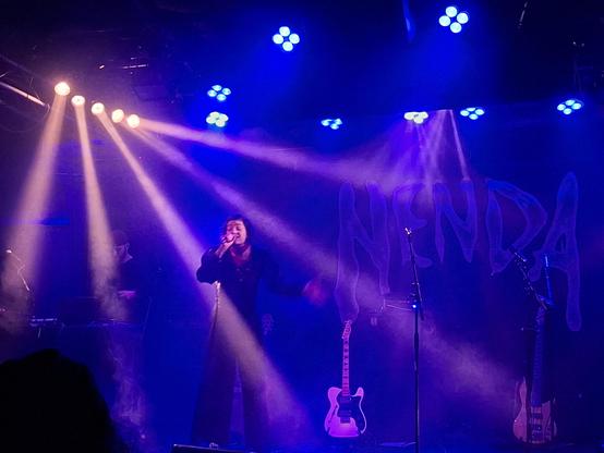 Foto von sirius.sue. Sie steht im blauen Licht mit vier hellen Lichtstrahlen. Sieträgt schwarze Kleidung und hält ein Mikrophon in der rechten hand