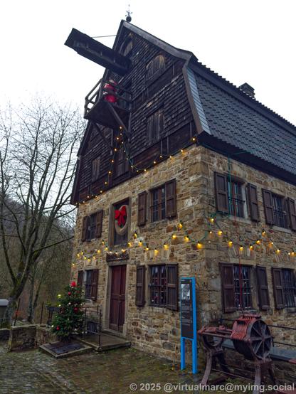A photo of a building with christmas decorations. There are multiple christmas lights hanging on the walls. A christmas tree is standing at the front. A christmas wreath with red ribbon is hanging over the door. A santa claus statue is standing on a balcony. It's still day and light.