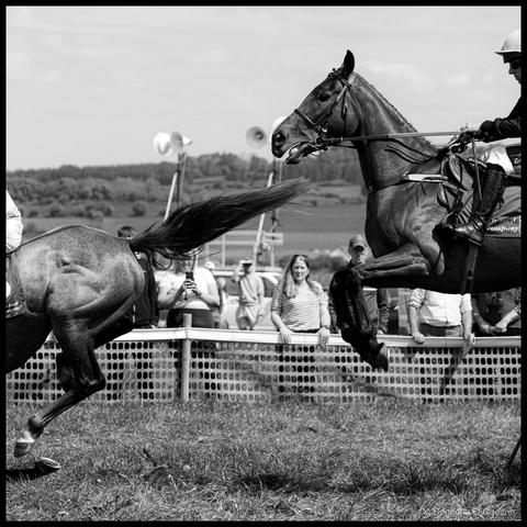 Black and white action photograph of a horse mid-jump with flowing mane and rider in dark clothing and white helmet sailing through the air over an out of photo jump at a point-to-point race, following another horse that has just landed. Blurred spectators in the background, countryside landscape and trees visible on the horizon, taken at Dawstown Point to Point.