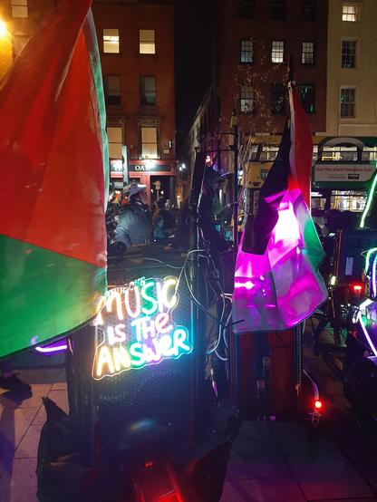 Photo of a large speaker sitting on a trailer attached to a bicycle. There is a colourful illuminated sign attached to the speaker which reads "Music is the Answer". There are large Palestinian flags attached to the speaker and the bicycle.