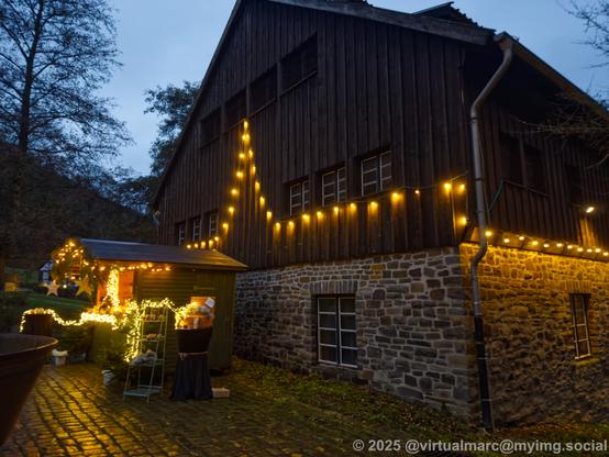 A photo of an old brick and wooden house (lower part are bricks, upper part is wood) with christmas lights hanging on the walls. A small shed from the christmas market is standing in front of it, which is full with christmas lights.