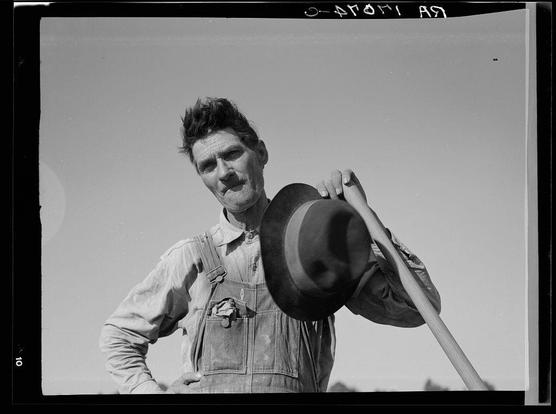 The image is a black and white photograph featuring an older man outdoors. He appears to be in his late 50s or early 60s, with spiky short hair and visible signs of aging such as wrinkles on the forehead and around the eyes. The individual has a rugged appearance, wearing overalls that are worn out from use, suggesting he may work manual labor.

He is holding a hat in his left hand and appears to be playfully pulling it off or showing it to someone outside of the frame with his right thumb extended outward as if making a dismissive gesture. His expression seems relaxed yet confident, possibly conveying a sense of pride or defiance.

The background is minimalistic but reveals that he stands against an open sky, hinting at a rural setting which aligns with historical context where such photos were taken to document the lives and struggles of American laborers during the Great Depression era in agricultural regions like Mississippi. The overalls have pockets on both front sides, possibly containing tools or personal items.

The image has text annotations suggesting it is part of a collection from White Laborer of the Mississippi Delta series by Dorothea Lange, which captures workers and their daily lives during challenging times. This specific photograph could be associated with Issaquena County in Mississippi as referenced in an URL provided alongside the photo link to source images on the website mentioned.

The black bac [...]