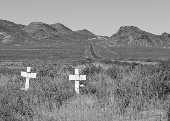 A black and white landscape photo of a pair of small white crosses. The crosses are stuck in the ground among various small desert shrubs next to an empty highway. The crosses are in the lower left of the frame. The highway cuts in at center right and disappears through a gap in distant hills. The sky is clear.