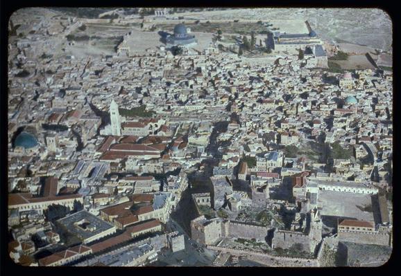 This image is an aerial view of the Old City in Jerusalem, showcasing a dense urban landscape with numerous buildings featuring red-tiled roofs. Prominent religious structures can be seen at various points across the cityscape, including domed and cruciform-shaped churches or mosques that stand out against the otherwise uniform architecture.

In the foreground, we observe historic stone walls enclosing what appears to be an ancient enclosure known as Herod's Fortress (or the Temple Mount), with a large open plaza within it. The streets seem narrow and winding among buildings closely packed together. Notable landmarks such as the Dome of the Rock and possibly other significant religious sites are visible in the distance, dominating the skyline.

The overall color palette consists mainly of earth tones—browns, reds, and tans—that blend with the natural surroundings. The photograph captures a historical snapshot of Jerusalem's Old City during a time when it was under different governance compared to today.
