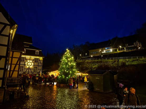 A photo of a cobblestone square. The cobblestone is still wet and reflecting the lights. A big decorated and shiny christmas tree is standing in the middle. Half timbered houses are on the left. They are decorated with christmas lights. The place is crowded in the distance. On the right side a hill is going up a building. The walkway up the hill and the building are lit with lights.