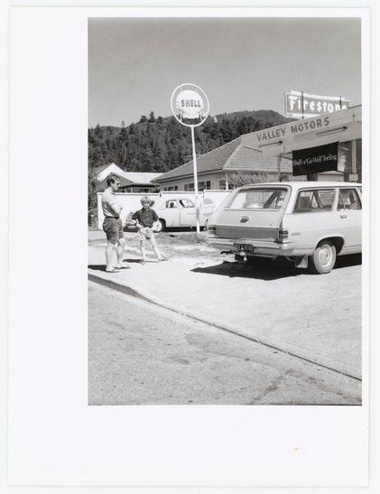 A black and white photograph captures a moment on an urban street. In the foreground, two individuals stand beside a parked vehicle labeled "VALLEY MOTORS." One person appears to be serving or interacting with another individual seated at what looks like a roadside service station counter, reminiscent of those found in mid-20th-century gas stations.

The backdrop features various commercial signs including one for Shell and Firestone tires. The environment suggests an era where such gas stations were common urban fixtures. There's also visible text on the side that is not fully legible due to perspective or distance but seems indicative of a service station setting with possible promotional material related to automobile maintenance.

The photograph carries a historical feel, likely from several decades past when roadside services and car culture played significant roles in daily life. The image evokes nostalgia for simpler times before modern fast-food establishments became ubiquitous on such stations' grounds.

Additional information about this image can be found within the context provided: Hutt Valley - Automobile service stations