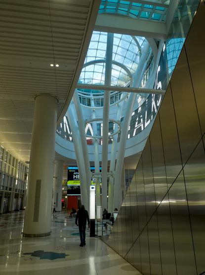 Looking up at a glass dome inside Salesforce Transit Center, beneath an escalator. The curvy wall beneath the dome is covered by a screen showing  large Latin letters.