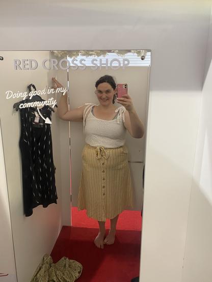 A person is standing in a mirror at a Red Cross shop, wearing a white top with straps and a yellow striped skirt. They are smiling and taking a selfie, with a few clothing items visible hanging nearby. The background features a sign with the text