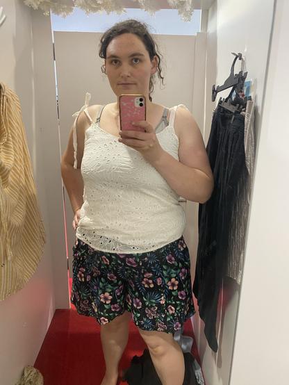 A person stands in a fitting room, wearing a white sleeveless top and black floral shorts. The mirror reflects the outfit and the colorful background. Clothing items hang nearby, and the floor is covered in red carpet.