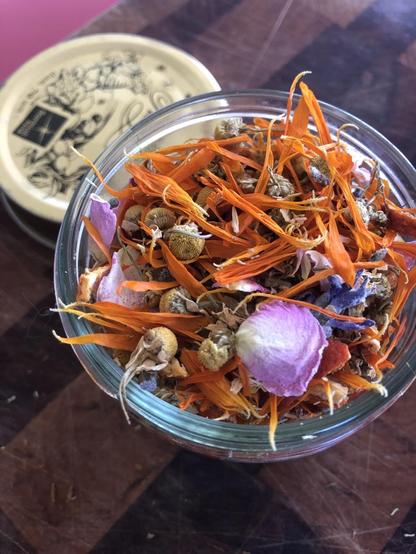 The tea in a glass jar with the gold lid behind. Mostly orange calendula petals, chamomile and white and pink rose petals are visible.