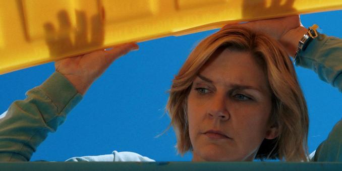 Carol (Rhea Seehorn) from Pluribus reaches overhead to open blue recycling bin lid, wearing green shirt, shot from low angle in moment from Episode 5