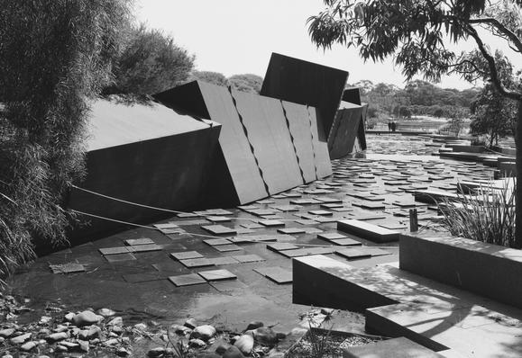 Black and white photograph of a flowing water feature stretching from the foreground and disappearing in the distance. In the flowing stream there is a grid of square paving stones at different heights, some above the water level and some below, giving a stepping stone effect. On the far side, there are large irregular shaped blocks.