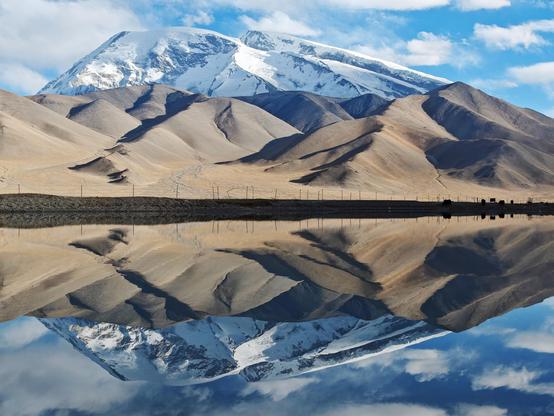 Muztagh Ata -"Father of Ice Mountain", Kunlun Mountains range, located in Western China (Xinjiang). Situated on the edge of the Tibetan Plateau and overlooks the Karakoram Highway. The mountain's summit is approximately 7,546 meters (24,757 feet) above sea level. The photograph beautifully captures the mountain's reflection in a still body of water, likely Karakul Lake (or a similar high-altitude lake or pond nearby