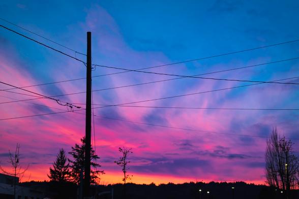 A photo of a utility pole with power lines silhouetted against a vibrant sunset sky. The sky transitions from deep pink and coral near the horizon through purple and lavender to rich blue above. Bare winter trees frame the composition on both sides with buildings and distant lights on in the dark hills at the bottom.