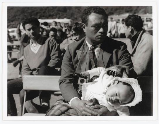 The photo is a black and white photograph depicting an outdoor gathering. In the foreground, there's a man dressed in a suit with a tie, sitting on what appears to be wooden bleachers or chairs. He holds a baby wrapped in a white garment, including a hat with ear flaps. The infant seems comfortable and secure against his chest.

The focus is slightly blurred around him, which highlights the subjects as they are meant for attention. Behind them, we see other attendees who appear engaged in conversations. Some wear sweaters over casual clothing while others have jackets on. Their expressions vary from attentive to neutral. In the background, a landscape with hills and some vegetation can be seen.

There is no direct indication of an event name or specific location beyond this being outdoors during daylight hours. It seems like people gathered for something significant given their attire but not in formal wear suggesting it could be a social function rather than ceremonial. The presence of both adults and children indicates family-friendly attendance to whatever they are attending, potentially hinting at community-oriented events such as festivals or local celebrations.

Without context provided by the photo's caption within Wanganui - Marae, there is no explicit indication about what this gathering might be for beyond these observations.