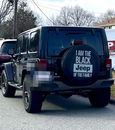A picture of the back of a black Jeep Cherokee that has a black spare tire cover. In white letters on this cover it says  I AM THE BLACK Jeep OF THE FAMILY!