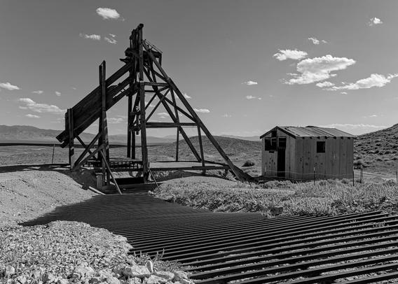 A black and white landscape photo of old mining equipment. Pictured are a wood "A" frame like structure used to winch loads of ore from a deep mine shaft. The "head frame" is upper center left. At center-right is a small single-room metal shack. From the bottom right to the base of the head frame are a series of parallel metal bars used to keep people and animals out of the mine shaft. They kind of look like road road leading up to the base of the head frame. The sky has a few small white puffy clouds.