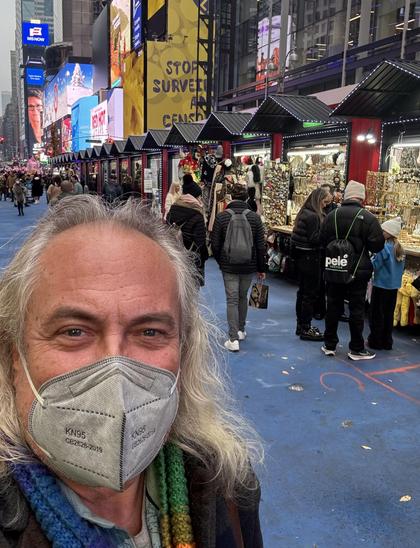 Selfie, me in a grey mask. Rows of holiday shops and the lights of Broadway behind.