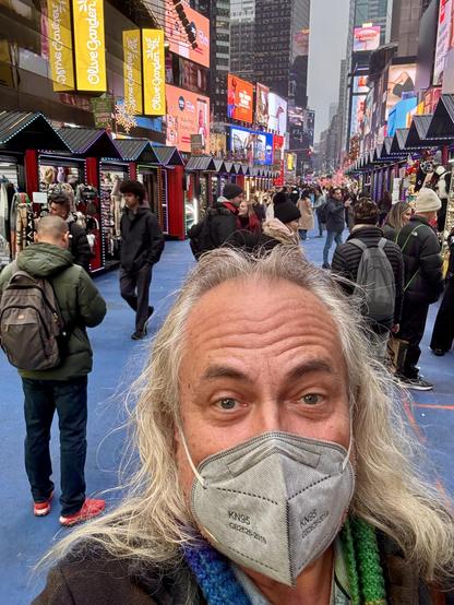 Selfie, me in a grey mask. Rows of holiday shops and the lights of Broadway behind.