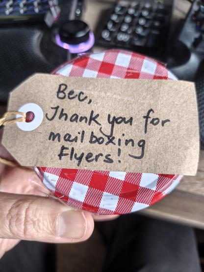 A closeup of the jar of jam that was in the letterbox. The lid has a red and white checkered pattern and taped to it is a brown paper label that says, Bec, Thank you for mailboxing flyers!