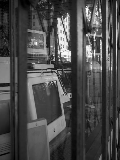 Black and white photo looking into a computer store that was closed up with metal gate. There're several vintage Macs inside, and a reflection of a high-rise building on the other side of the street