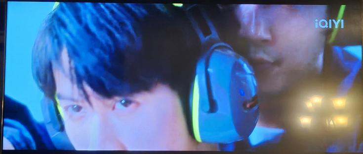 Paused TV with a close-up of two men wearing hearing protection, the taller one directly behind the shorter man, arms around him, to help him aim. Tan Jianci is the shorter man.