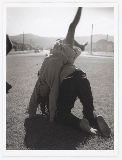 The black and white photograph depicts a dynamic scene of two individuals engaging in what appears to be a playful or acrobatic maneuver on grass. The person lying flat on the ground is wearing trousers with visible seams, suggesting practical clothing suited for activity. Above them, another individual is upside down above their feet, balancing themselves by wrapping their legs around and holding onto the first person's body; this creates an arc-like shape in mid-air.

The backdrop features a rural setting with hills or low mountains under clear skies, along with some distant structures that might be buildings, indicating it could be near a residential area. The contrast of shadows on the grass suggests sunlight from above, casting defined shapes around the figures and objects present.

There's an element of movement captured in this still image as evidenced by the extended leg reaching towards the sky which adds to the sense of motion within the composition. It evokes themes such as childhood playfulness, physical strength, balance, or even a cultural practice. The absence of color highlights contrasts between light and shadow, emphasizing textures like grass blades against smooth skin tones.

The photograph has an element of candidness that suggests it was taken spontaneously without staging, capturing a moment in time rather than a posed shot intended for artistic display.