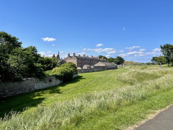 A row of historic stone buildings with multiple chimneys stretches along a low defensive wall, bordered by wide, neatly cut grass and taller, wilder growth near the path. The scene is backed by scattered mature trees and a bright blue sky with a few small clouds. A paved walkway curves through the right side of the frame, where a few distant people are visible enjoying the open space. On the left, dense greenery and a stone wall partially conceal sections of the buildings, while the open grassland in the foreground gives a clear view of the structures and the low, rolling terrain beyond.