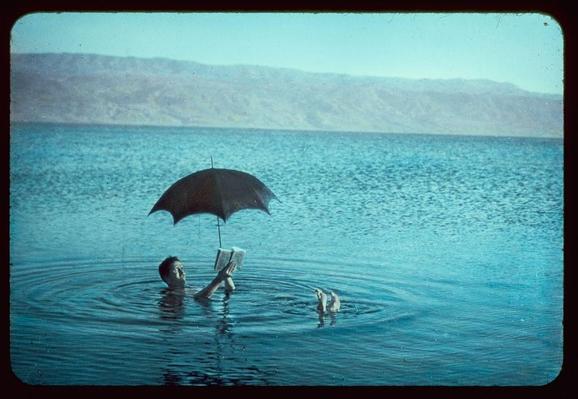 A person is floating on their back in a large body of water, supported by the buoyancy. They are holding an open book and using it as shade with a black umbrella. The calm blue water reflects sunlight, indicating clear weather conditions. In the foreground, another individual or object floats nearby at a shallower depth than the main person. The backdrop features a hilly landscape under a sky that appears to be partly cloudy but still bright enough for outdoor activities like reading in the water.